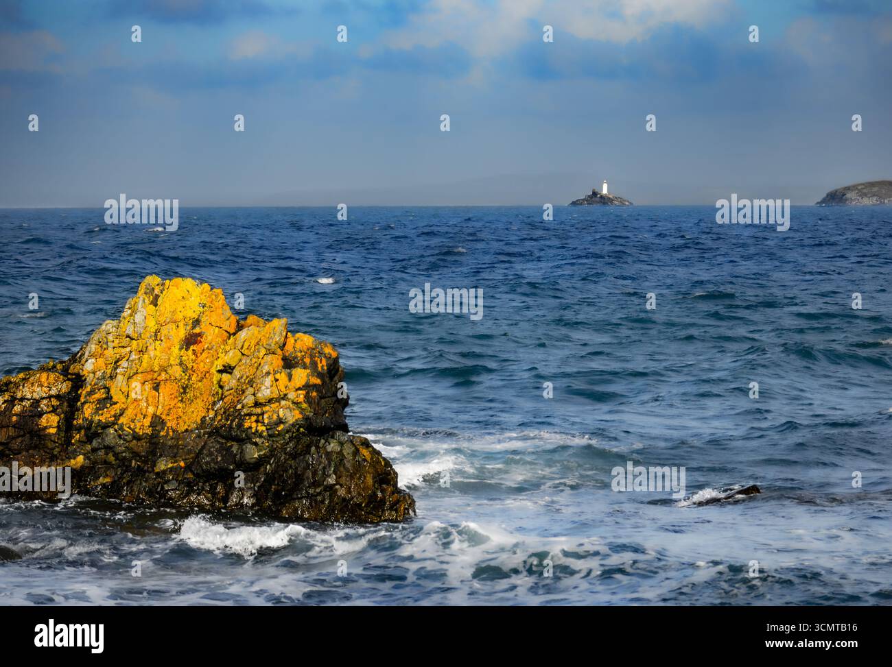 Il lichene dorato ha ricoperto rocce in primo piano con un faro su un'isola lontana attraverso le acque blu dell'oceano sotto il cielo nuvoloso sulla costa panoramica Foto Stock