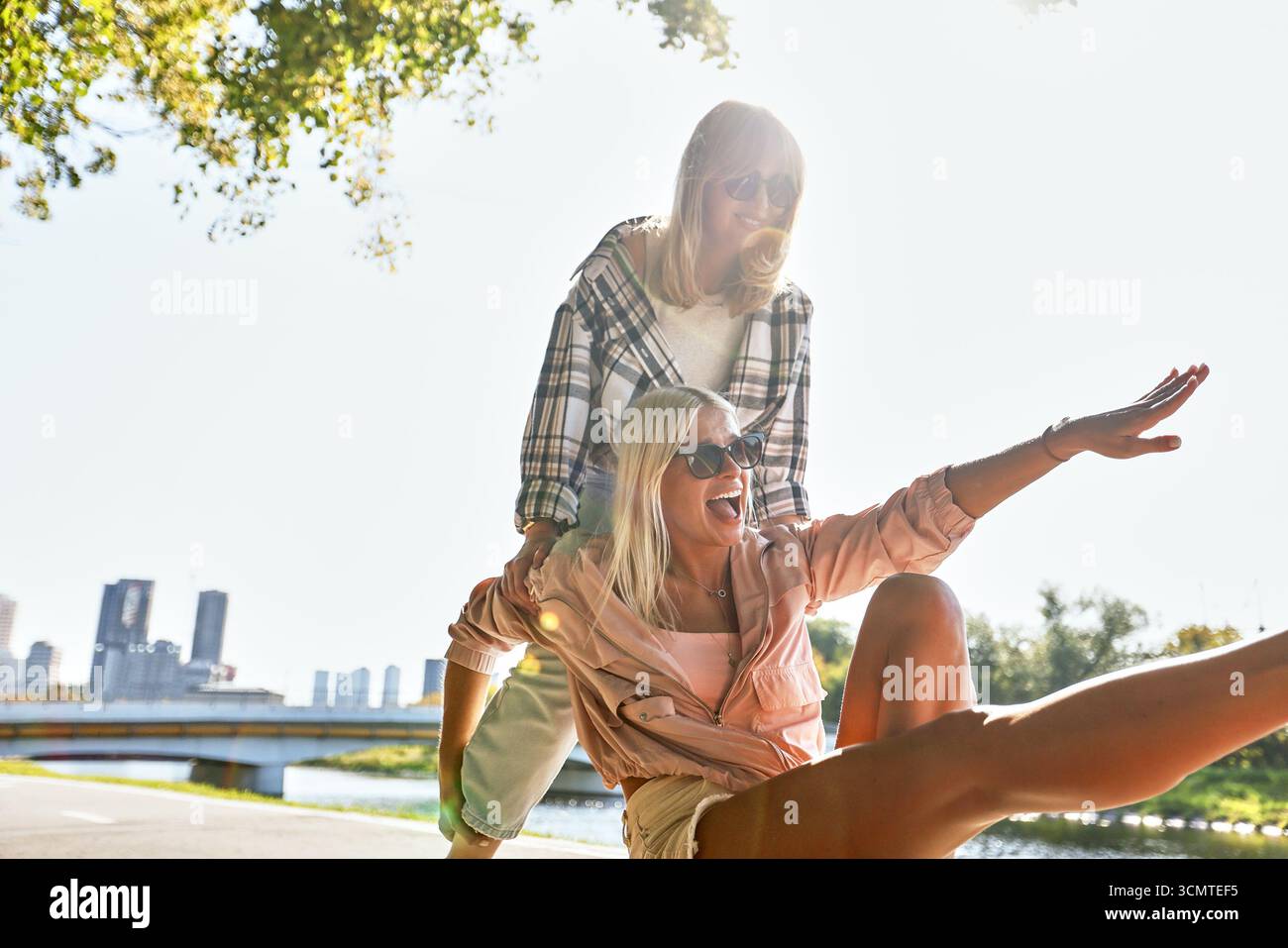 Due allegre donne vestite con abiti casual condividono un momento di gioia all'aperto. Sullo sfondo di alberi e di uno skyline cittadino, mostrano un'atmosfera spensierata Foto Stock
