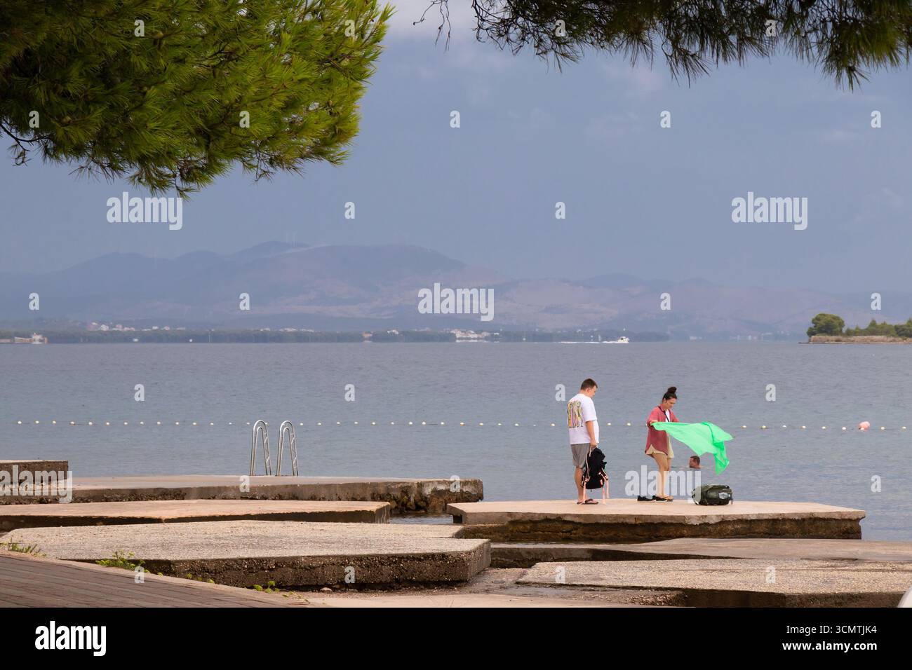 Vodice, Croazia - 30 agosto 2025: Giovane coppia che arriva su una spiaggia vuota in una giornata estiva con tempo variabile su una costa Foto Stock