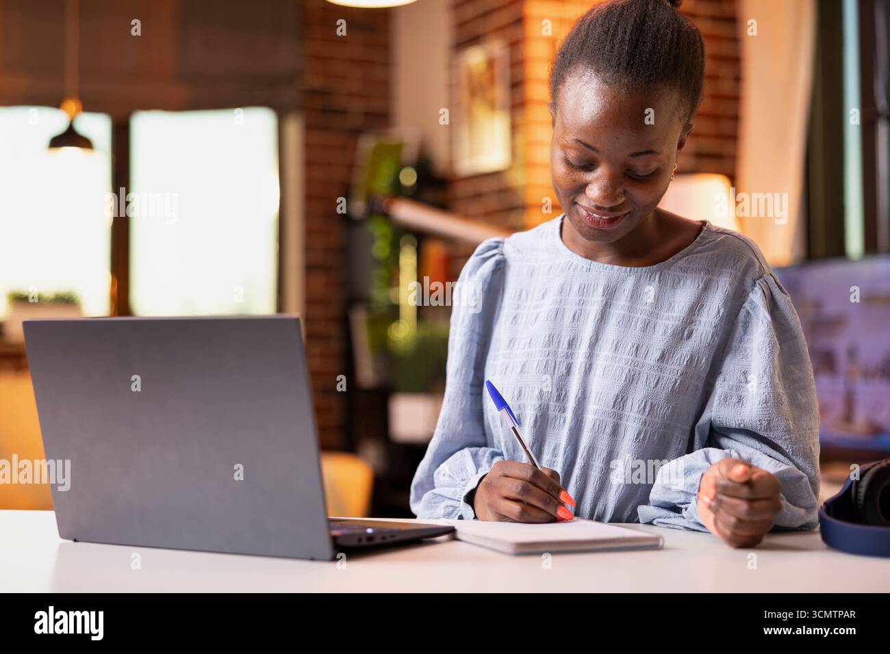 Lavoratrice freelance concentrata che lavora in interni, esamina le prestazioni di vendita dei notebook e prende appunti. Lavoratore remoto che scrive idee in blocco note mentre gestisce le attività di reporting e organizzative. Foto Stock