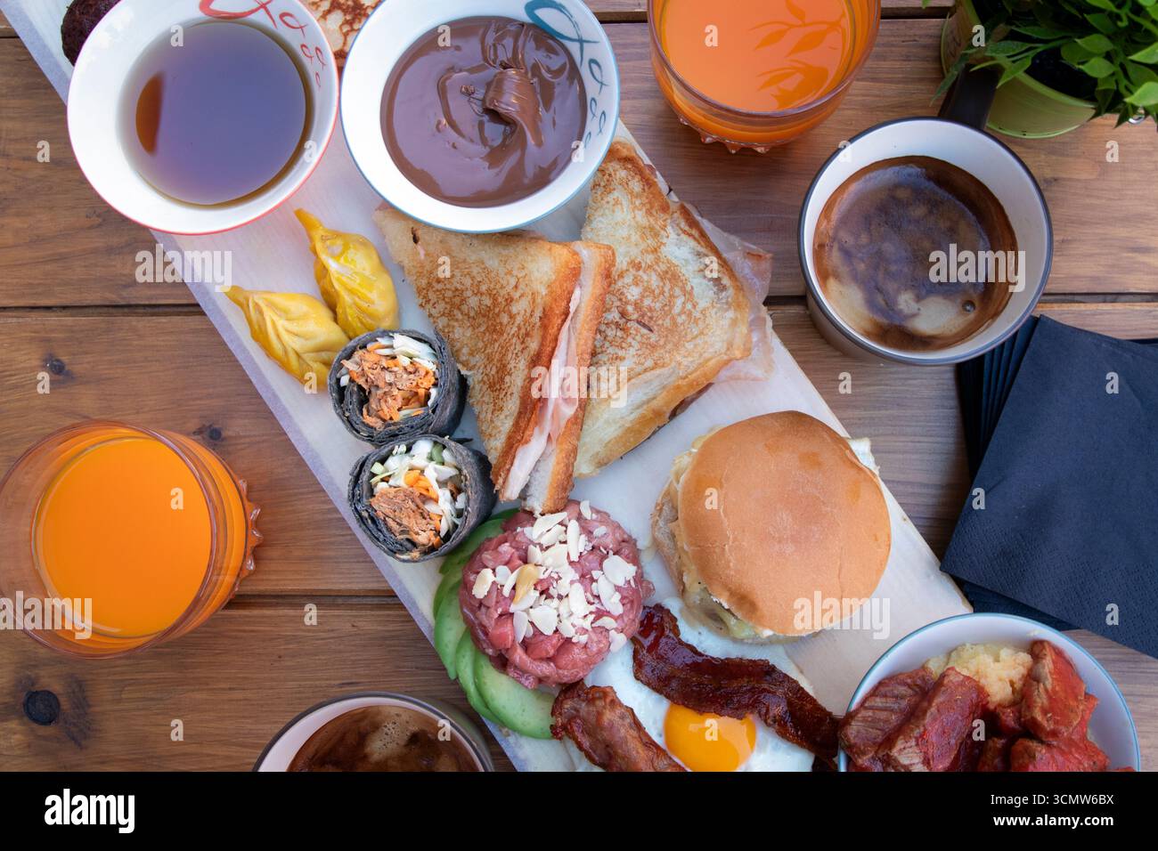 Vivaci shot di brunch condiviso con pancake, sushi, uova, hamburger e bevande. Composizione artistica e atmosfera conviviale su tavola rustica. Foto Stock
