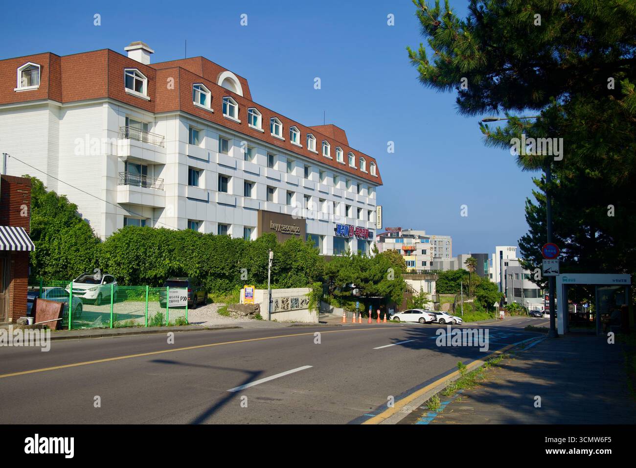 L'hotel Heyy Seogwipo si affaccia su un viale in salita fiancheggiato da alberi e piccoli negozi ai margini del centro cittadino, mostrando il basso paesaggio urbano. Foto Stock