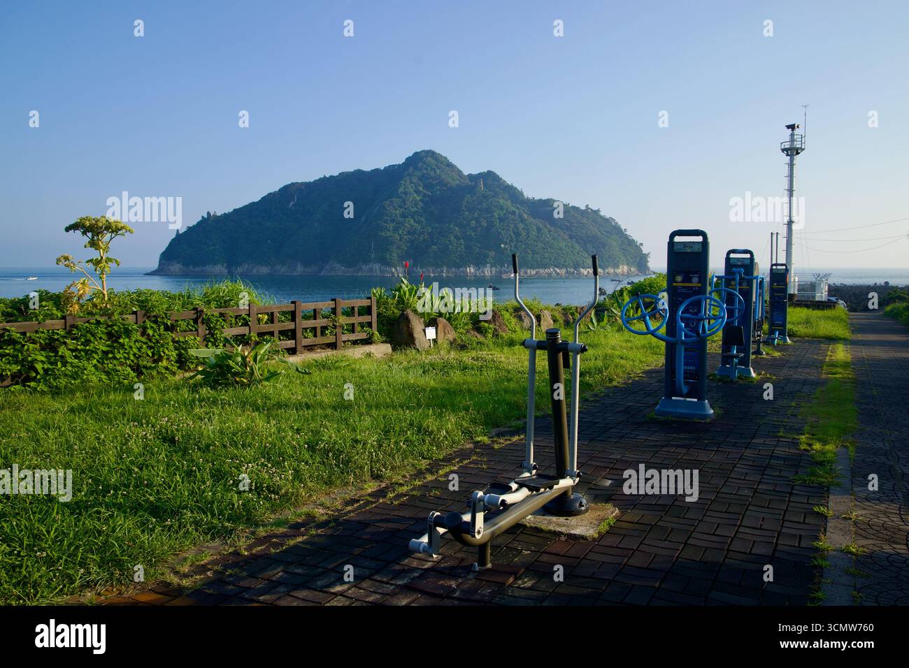 Le macchine per il fitness all'aperto si trovano lungo un sentiero sul mare a Seogwipo con Munseom che sorge appena al largo, visto oltre una recinzione bassa, un vergine erboso e un piccolo ettaro Foto Stock
