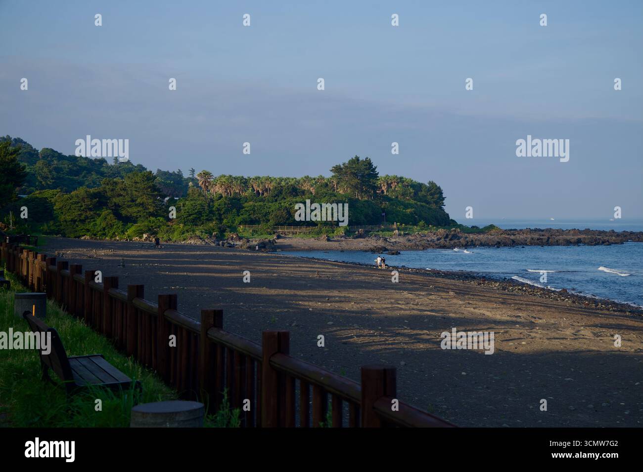 La sabbia nera della spiaggia di Hahyo Soesokkak si estende verso un punto roccioso e un promontorio ricoperto di palme, mentre piccole onde lambiscono le pietre alla bocca del so Foto Stock
