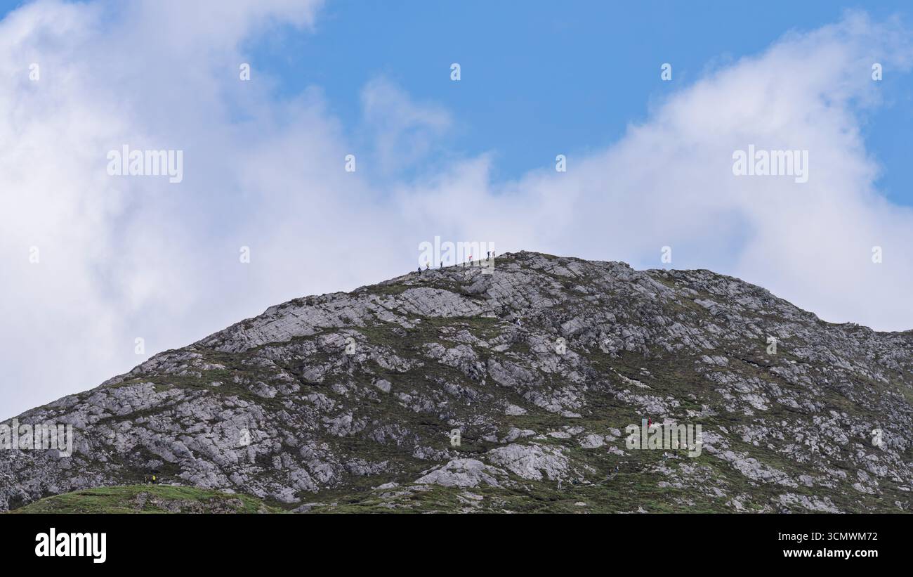 Camminatori distanti sulla cresta rocciosa della cima di Diamond Hill sotto una nuvola patinata, Connemara National Park, County Galway, Irlanda, visti dal sentiero. Foto Stock