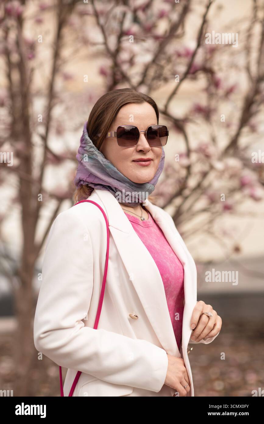 Donna Magnolia Fiori Primavera donna in sciarpa, occhiali da sole, blazer bianco posa vicino a magnolia albero in primavera per la moda. Foto Stock