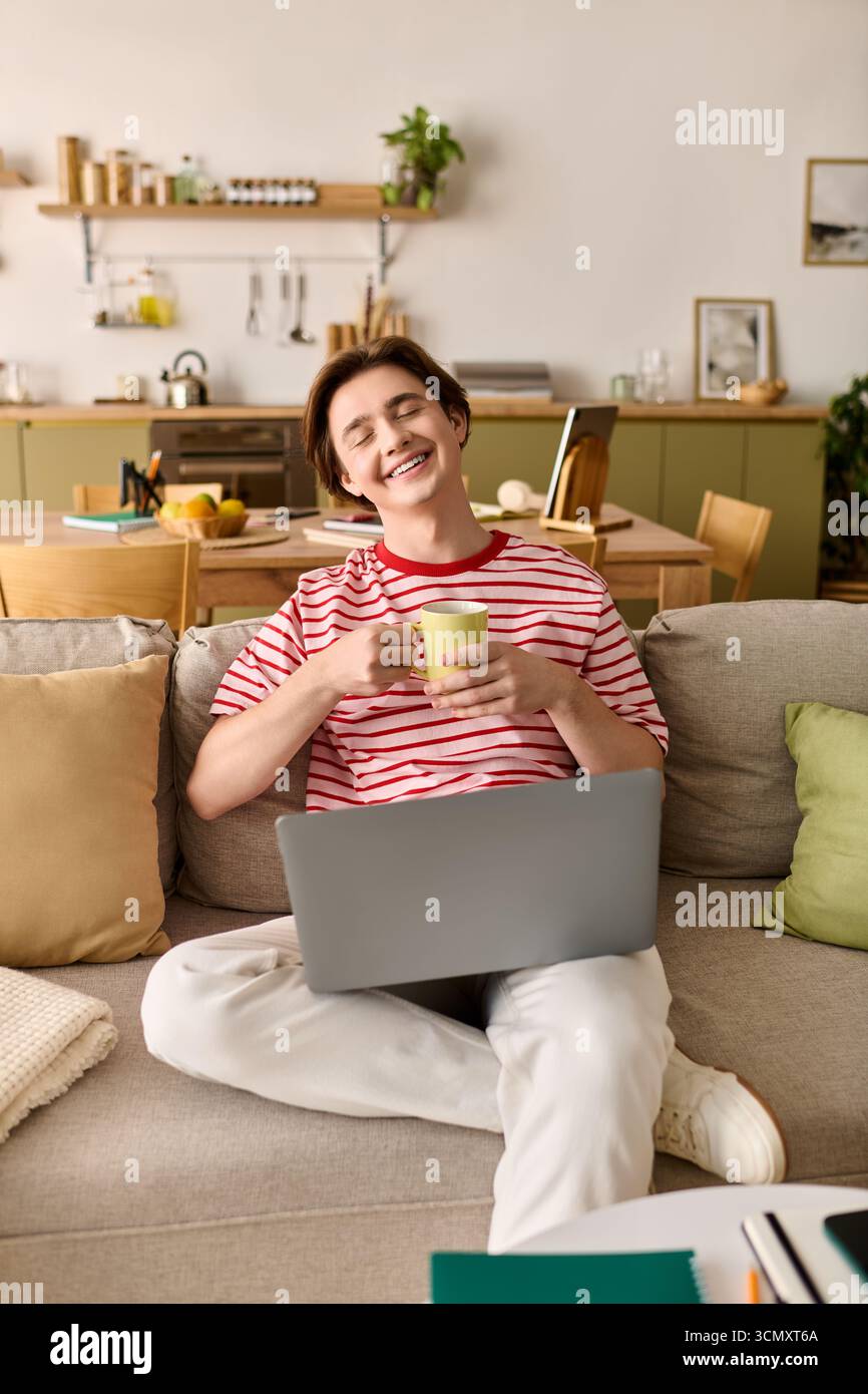 Un giovane uomo si rilassa su un divano mentre studia online con un portatile e beve in mano. Foto Stock