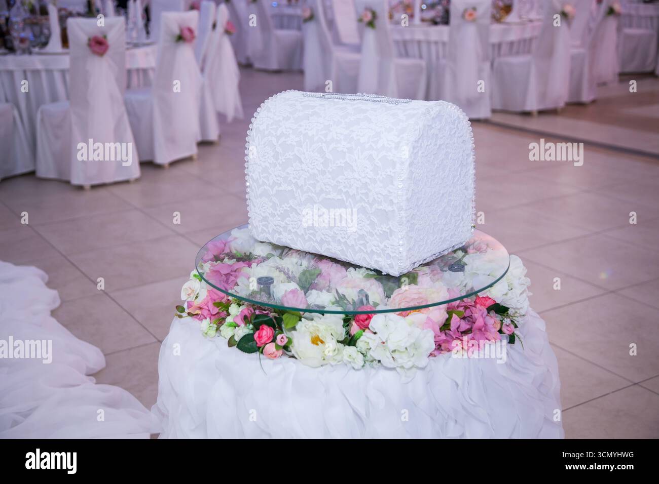 Un portamonete bianco splendidamente realizzato poggia su un piedistallo decorato con fiori in un luogo di ricevimento. Morbide decorazioni floreali circondano la scatola, enhan Foto Stock