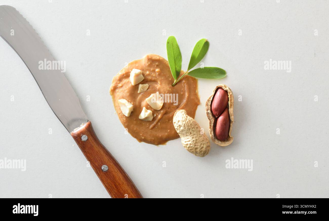 Macro fondo concettuale di burro di arachidi con pezzi su un tavolo bianco con coltello e arachidi che mostrano i chicchi, decorati con foglie verdi. Superiore Foto Stock