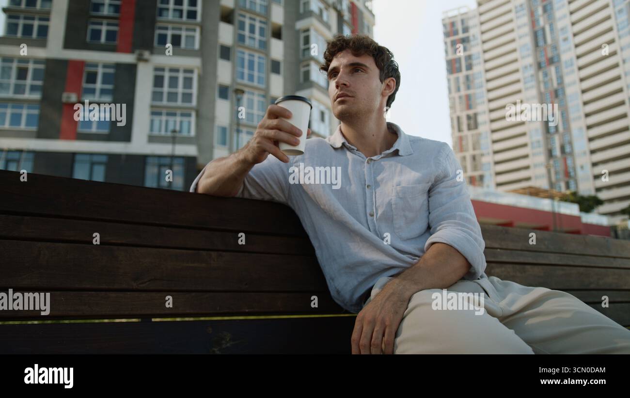 Uomo europeo caucasico seduto sulla panchina maschio che tiene tazza pausa caffè bere takeaway ordine cliente acquirente rilassarsi gustoso drink Guy studente uomo d'affari Foto Stock