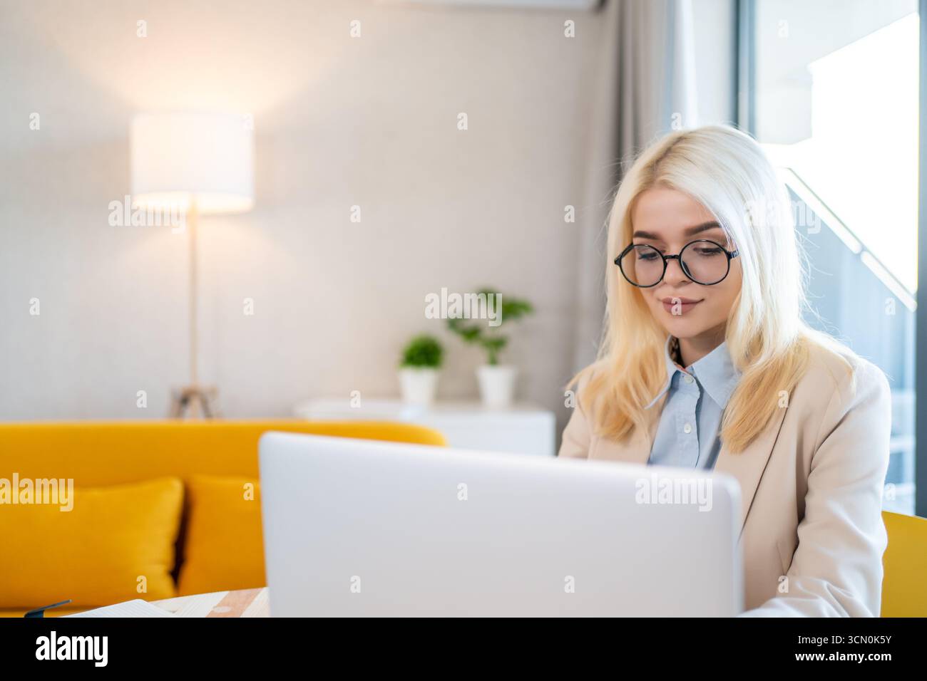 Donna d'affari bionda con occhiali che utilizza un computer portatile in un ambiente moderno e accogliente per lavorare in remoto, lavorare in freelance, ufficio domestico, comunicazione digitale, successo professionale, formazione online, produttività. Foto Stock