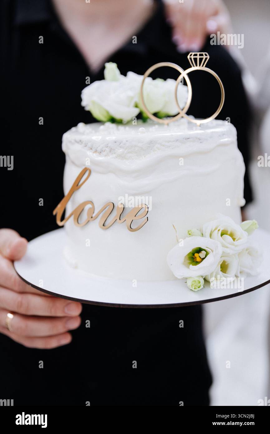 Elegante torta nuziale con anelli e fiori bianchi, tenuta dagli sposi in occasione della celebrazione Foto Stock