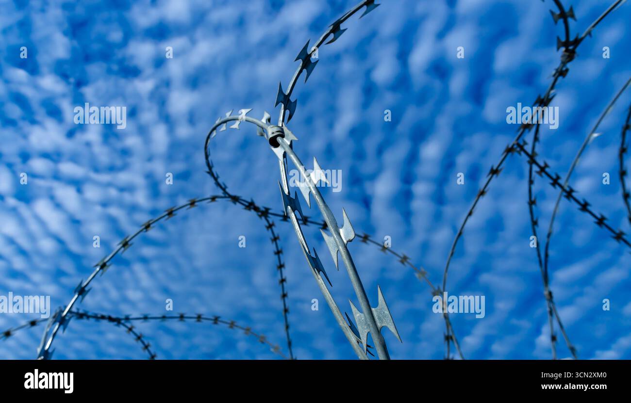 Gli anelli in filo spinato in stile NATO si stagliano contro un cielo blu con nuvole, simboleggiando sicurezza, controllo e confini. Foto Stock