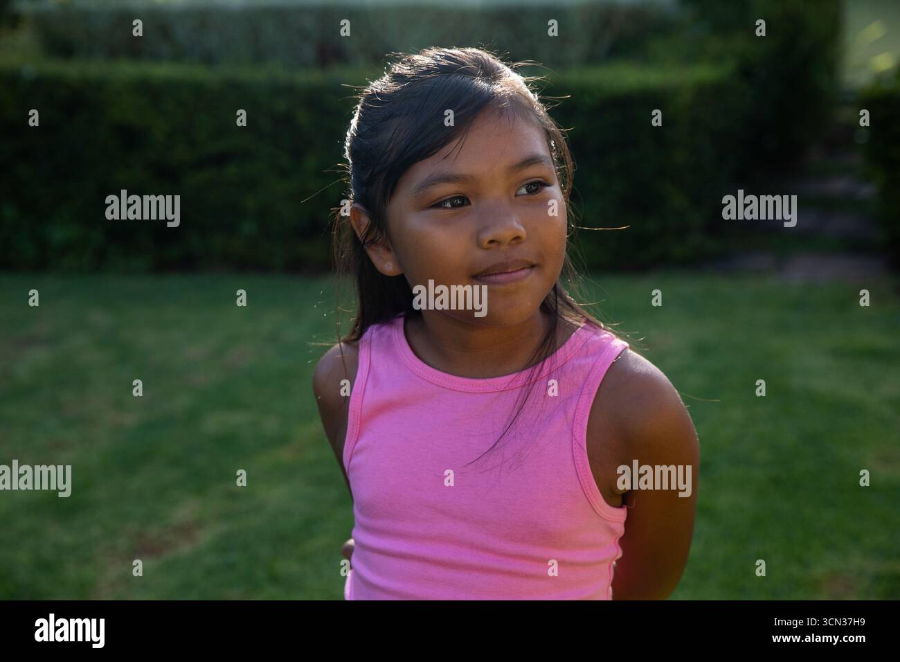 Ragazza asiatica in piedi con cura nel giardino sul retro con la parte superiore rosa accanto alla siepe rifilata Foto Stock