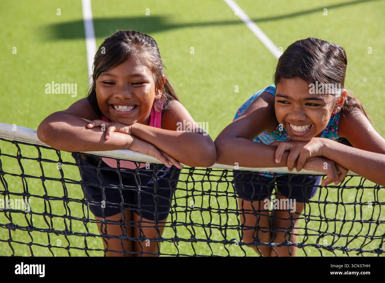 Bambine asiatiche che si appoggiano su una rete da tennis su un campo di erba verde con linee bianche sotto la luce del sole Foto Stock