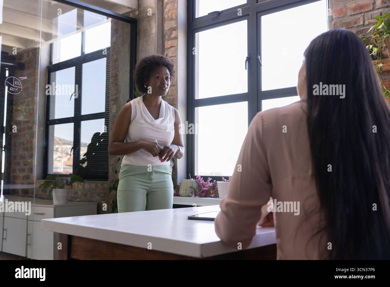 Diversi colleghi che parlano vicino alla finestra nell'ufficio del loft alla scrivania con piante in vaso per tablet Foto Stock