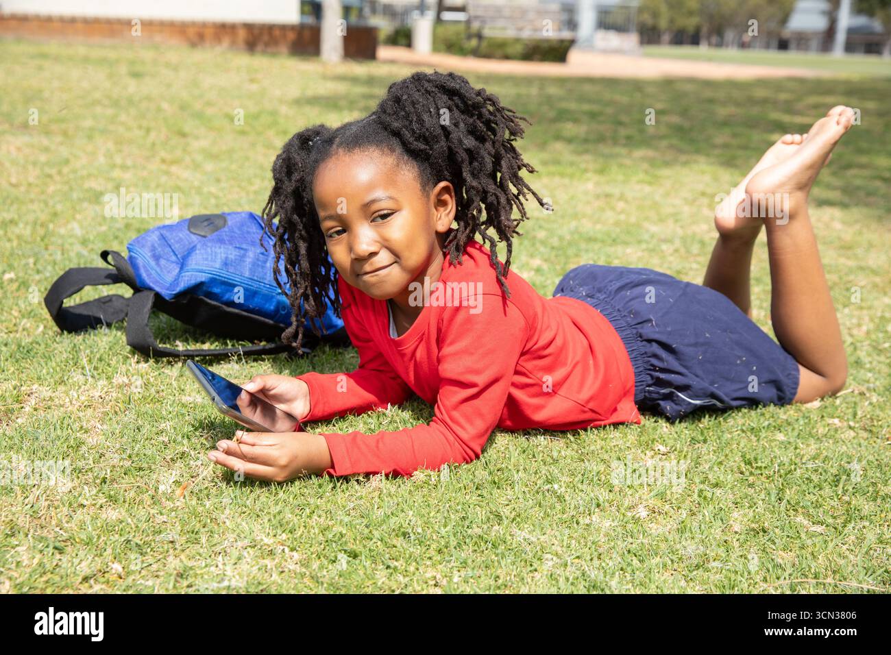 Bambino afroamericano in cima rossa disteso sul prato del parco con smartphone con zaino Foto Stock