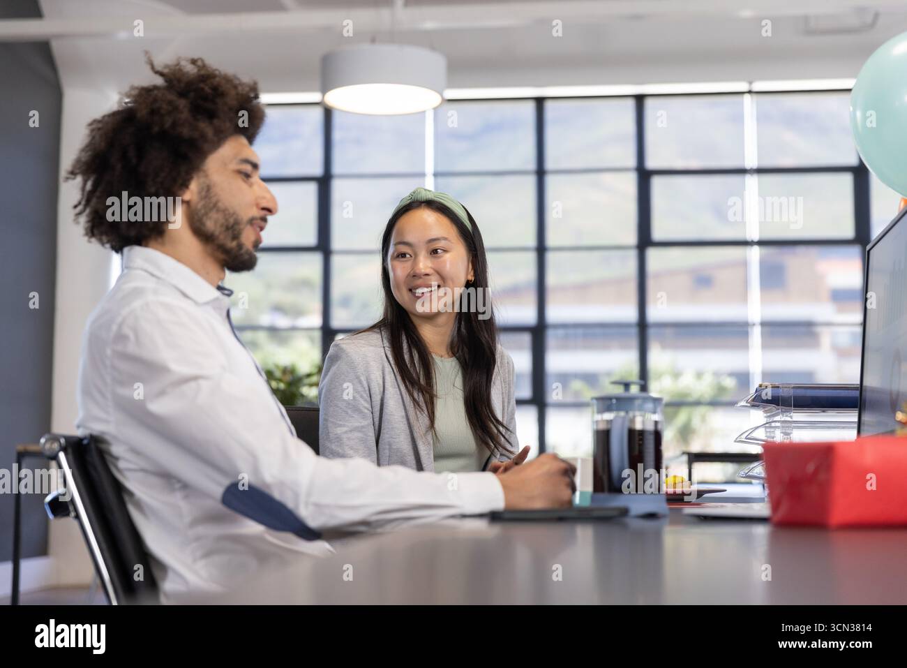 Colleghi diversi che discutono di piani mentre sono seduti alla scrivania dell'ufficio con computer, scatola regalo, palloncino Foto Stock