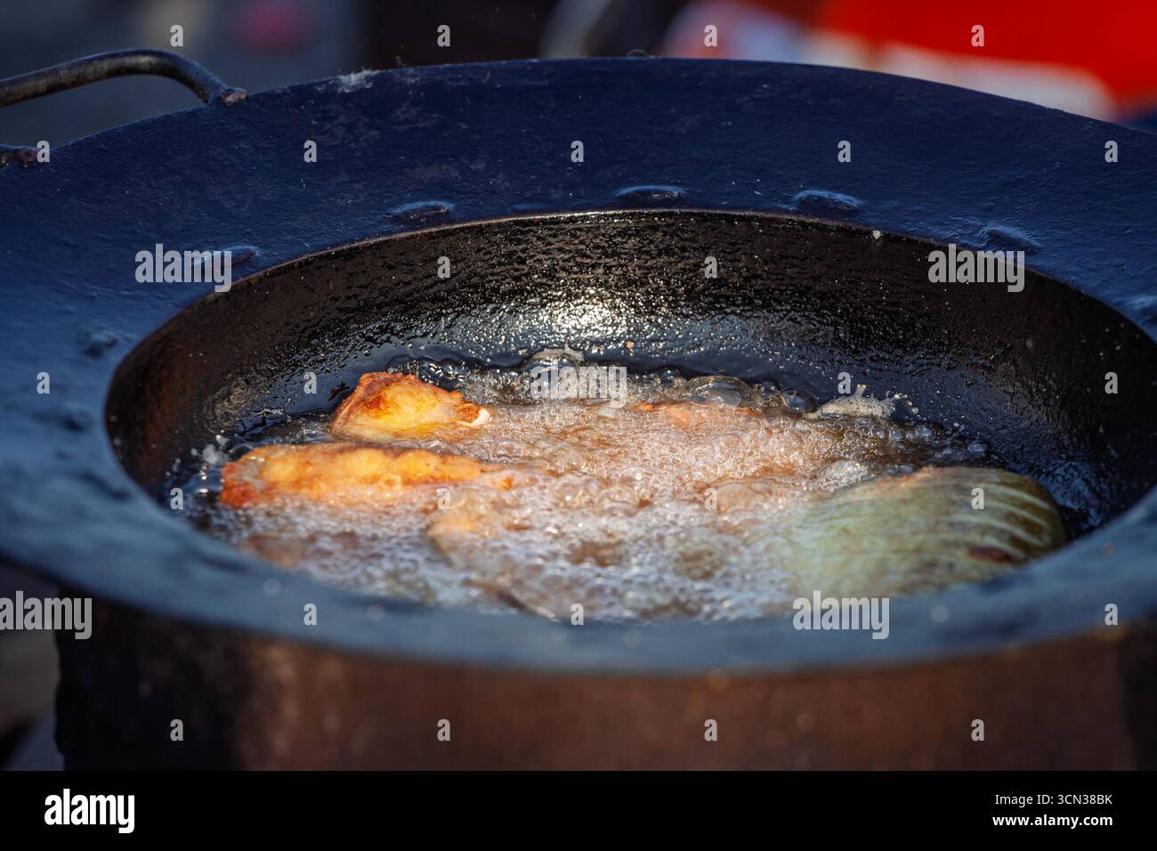 Vista ravvicinata dei pezzi di pesce fritti in olio caldo come parte della preparazione della zuppa di pesce serba (riblja corba). Olio gorgogliante, bordi dorati e una coppa in ghisa mostrano t Foto Stock