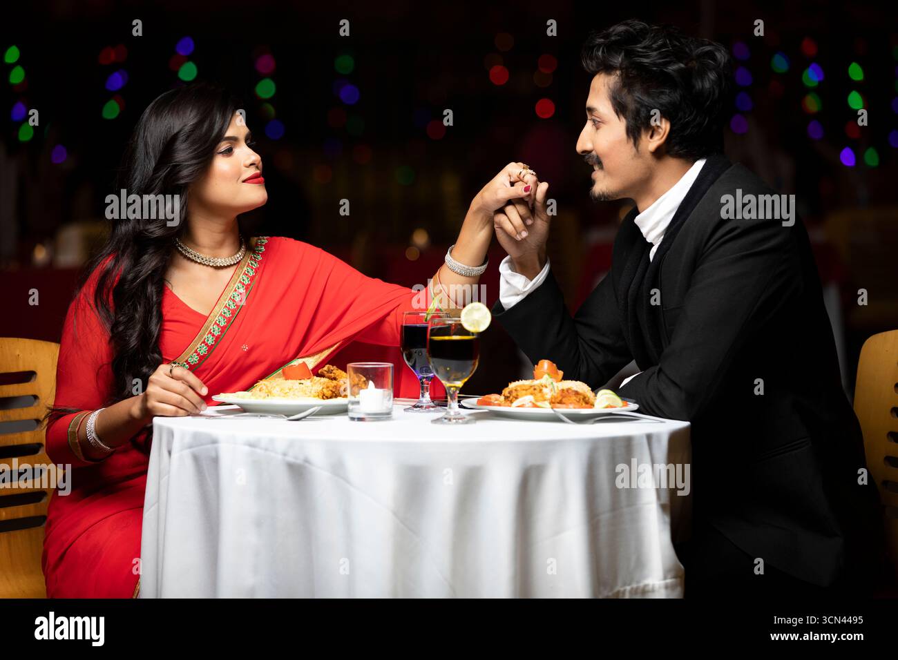 Una bella coppia in abbigliamento tradizionale che si tiene per mano e sorride l'un l'altro durante una cena romantica in un ristorante con luci calde nella Foto Stock