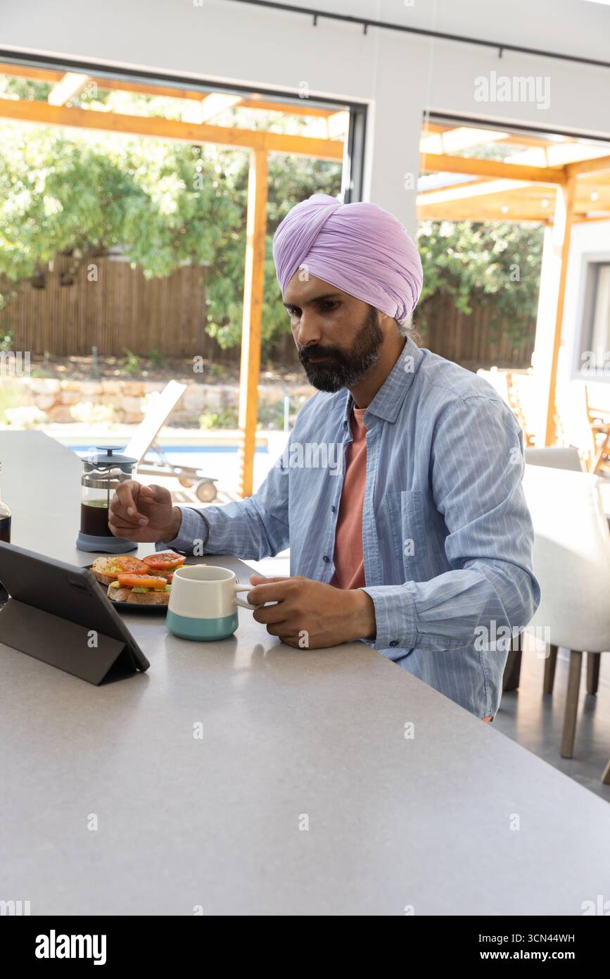 Un uomo indiano seduto al piano di lavoro sorseggiando un caffè dalla stampa francese mentre usa un tablet accanto ai bagel Foto Stock