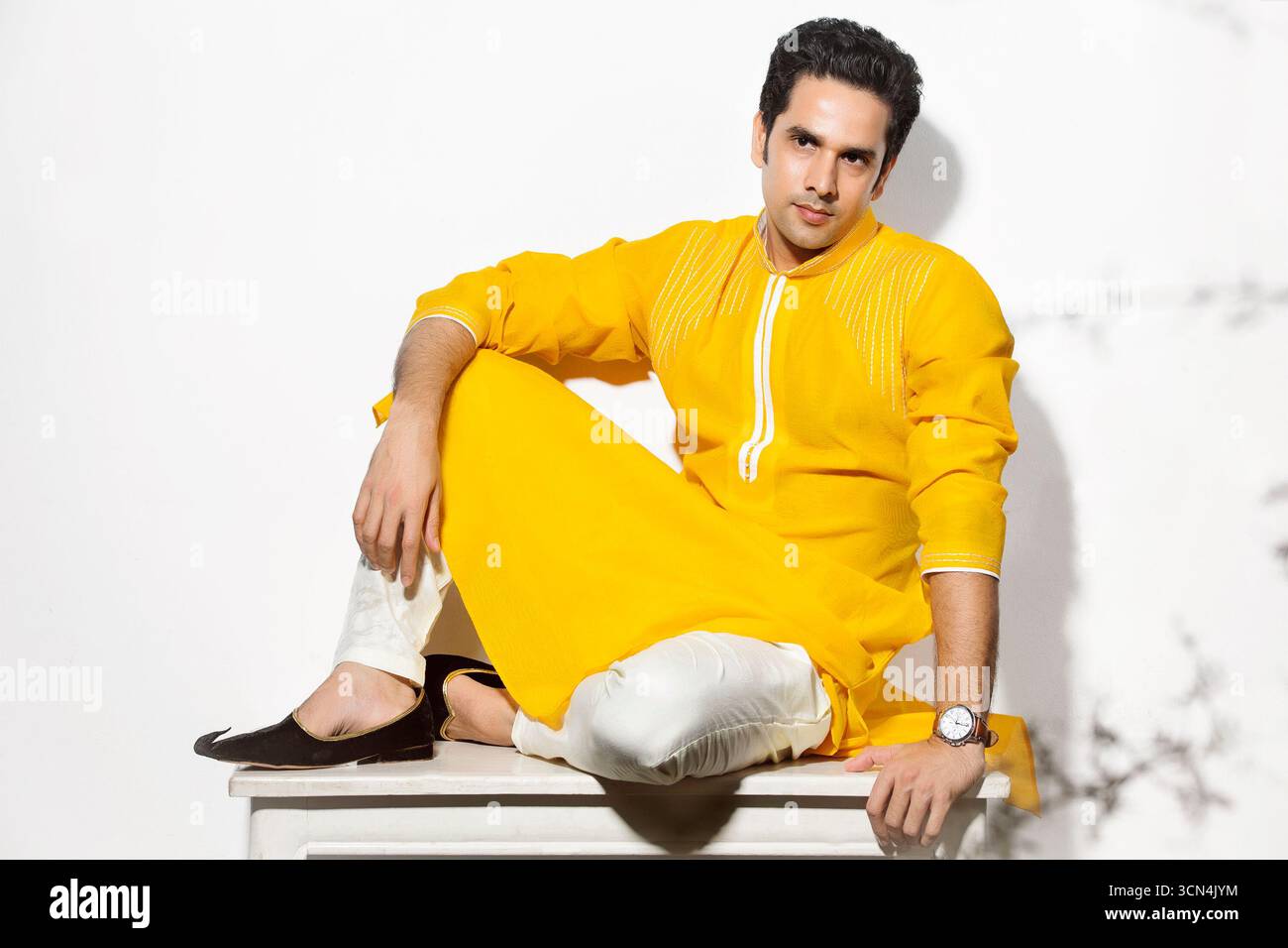 Un elegante uomo dell'Asia meridionale in un vivace kurta giallo e churidar bianco si posa con uno sguardo sicuro mentre si siede su uno sgabello in uno studio. Foto Stock