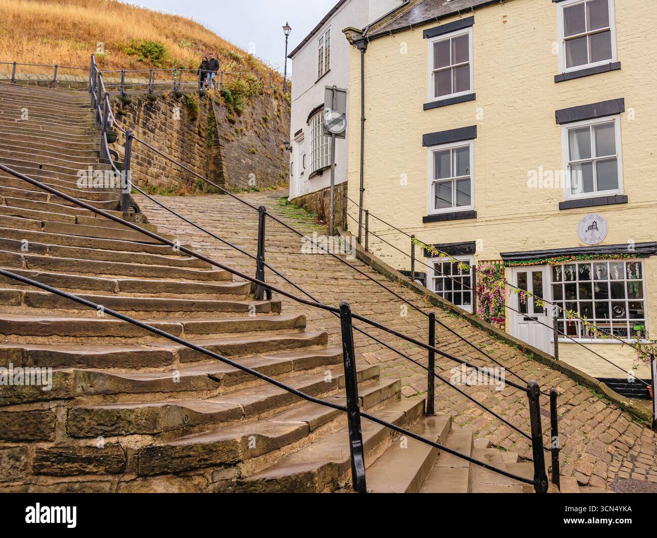 I famosi 199 gradini che conducono all'abbazia di Whitby. I gradini si curvano verso l'alto e oltrepassano un banco erboso. Una casa è la base con un cancello. Foto Stock