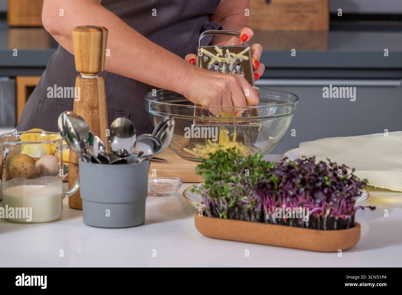 Chef donna che grattugia gli ingredienti freschi in una ciotola, con utensili da cucina ed erbe nelle vicinanze, creando un'atmosfera vivace per la preparazione culinaria Foto Stock