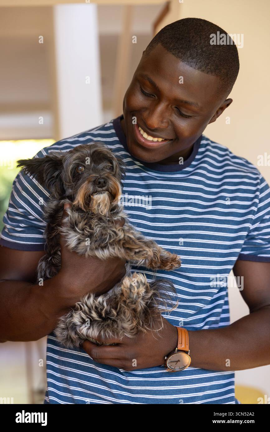 Uomo afroamericano con un piccolo cane soffice contro il petto che indossa un orologio in pelle nel corridoio di casa Foto Stock