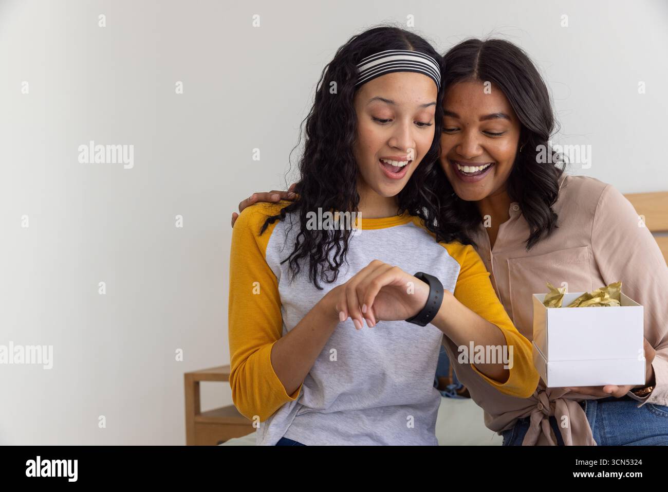 Mamma e figlia adolescente diverse sedute sulla panchina in camera da letto a esaminare smartwatch e confezione regalo Foto Stock