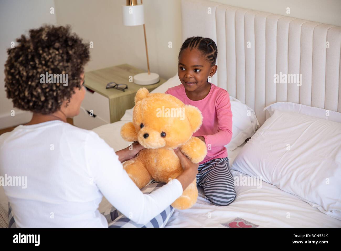 Sorelle afroamericane che consegnano peluche sul letto con lampada e bicchieri sul comodino Foto Stock