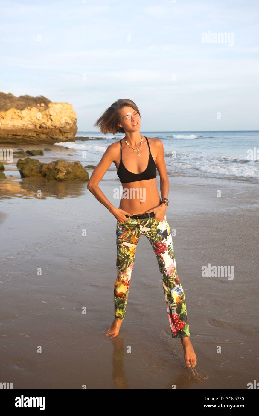 Bella donna sportiva naturale con pantaloni in fantasia floreale e reggiseno nero si gode la spiaggia del tramonto e la formazione rocciosa unica in Portogallo. Felice, allegro Foto Stock