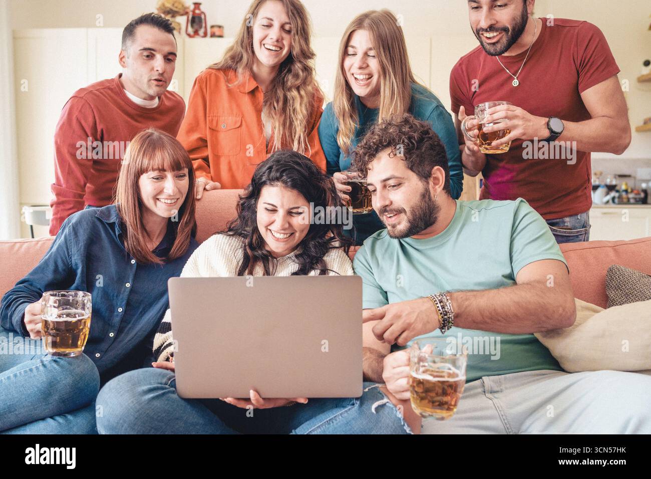 Un gruppo eterogeneo di giovani amici felici si riunisce intorno a un notebook su un divano, sorridendo e guardando un film o trasmettendo in streaming contenuti a casa. Foto Stock