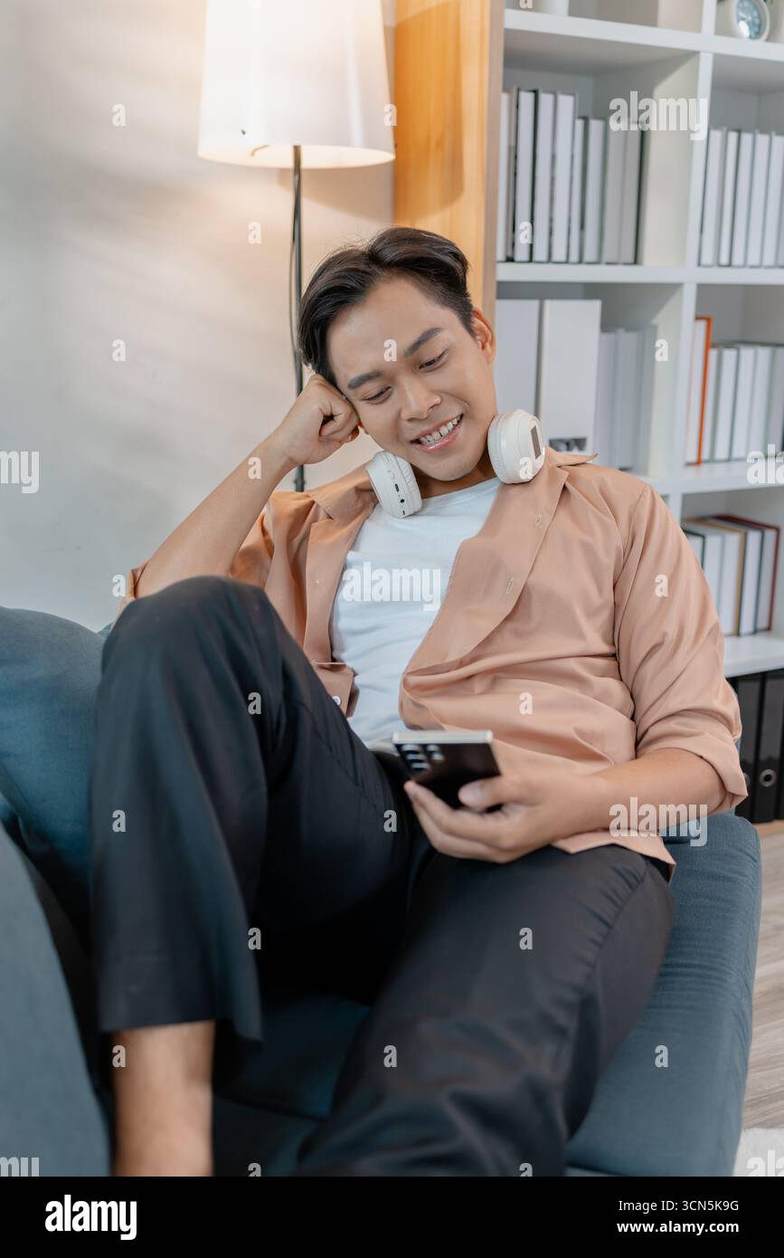 Un giovane uomo si rilassa a casa, sorridendo mentre usa il suo smartphone. Ama trascorrere il tempo libero con le cuffie intorno al collo, seduto comodamente e comodo Foto Stock
