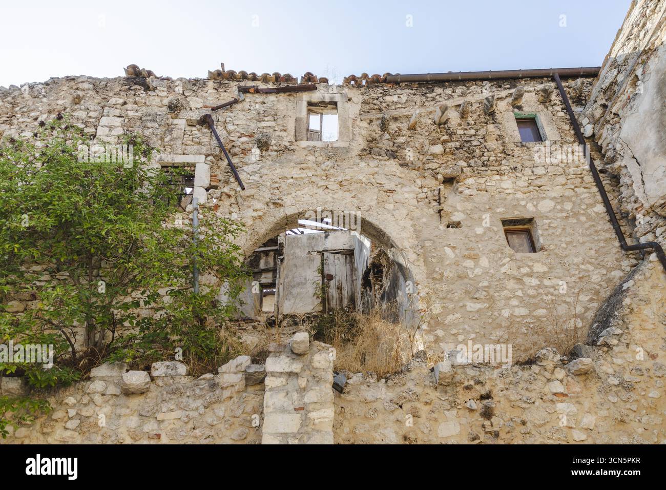 Veduta delle mura in pietra e della porta ad arco, scorci di spazi interni, che accennano alla storia e al degrado sotto un cielo blu, Castelvecchio Calvisio, Abruzzo, Italia. Foto Stock