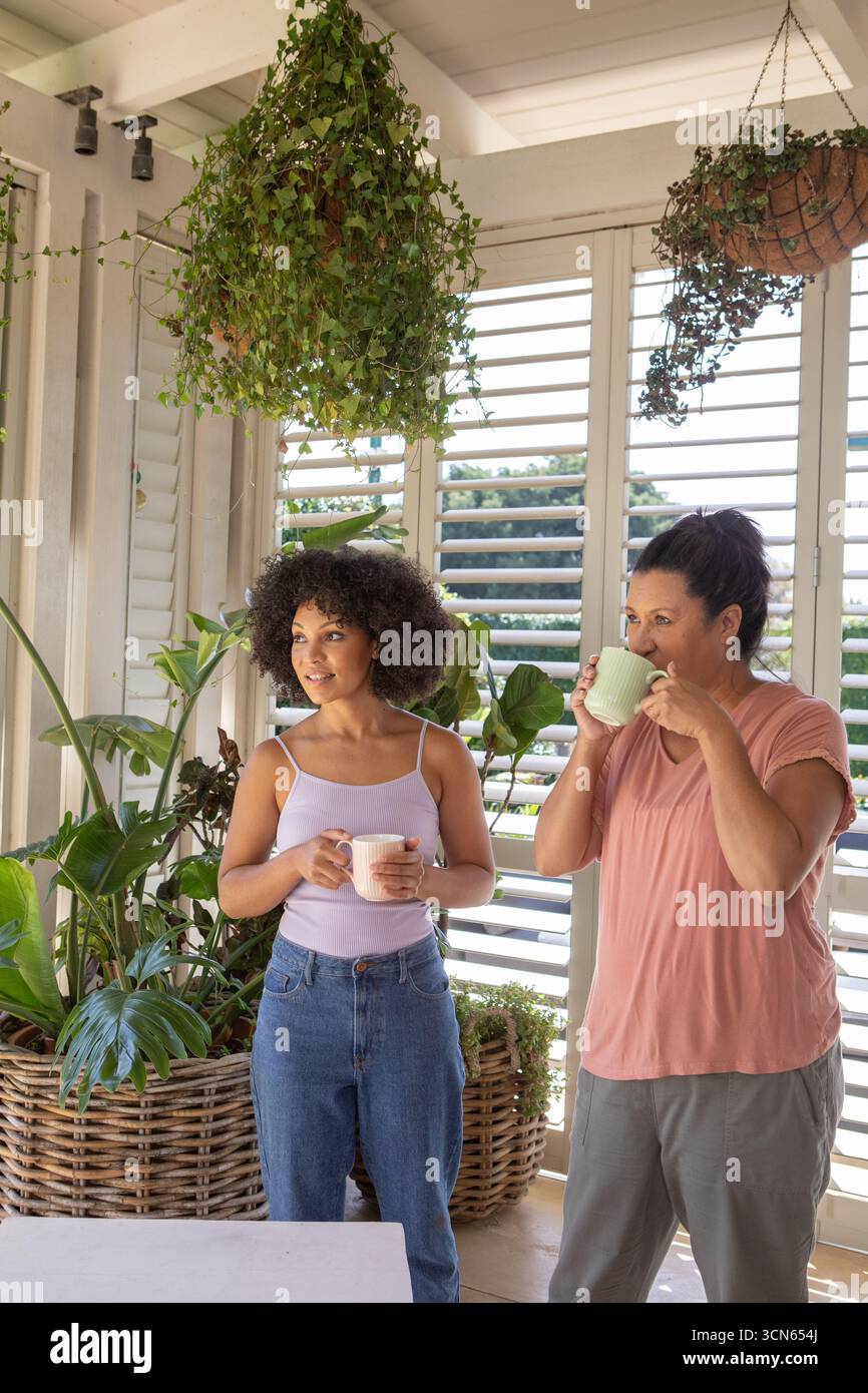 Madre e figlia in piedi su un portico coperto illuminato dal sole che tiene tazze in ceramica accanto a piante lussureggianti Foto Stock