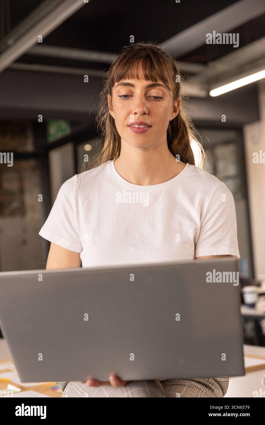 Donna che tiene un notebook e guarda lo schermo in ufficio con luci a LED e pareti divisorie in vetro Foto Stock