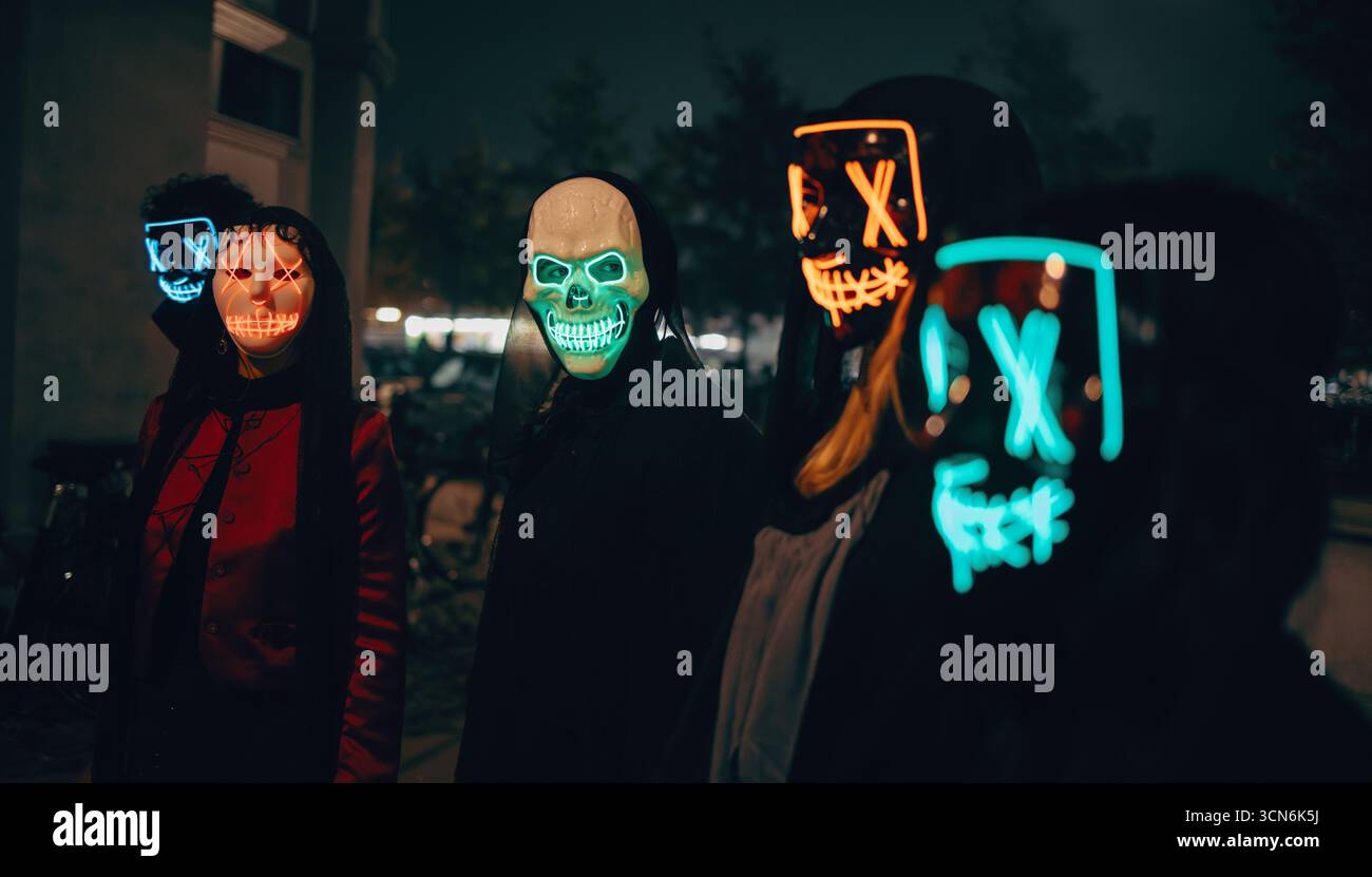 Cinque persone che indossano maschere al neon brillanti e abiti scuri si riuniscono all'aperto di notte, creando una vista misteriosa e sorprendente contro la b serale Foto Stock