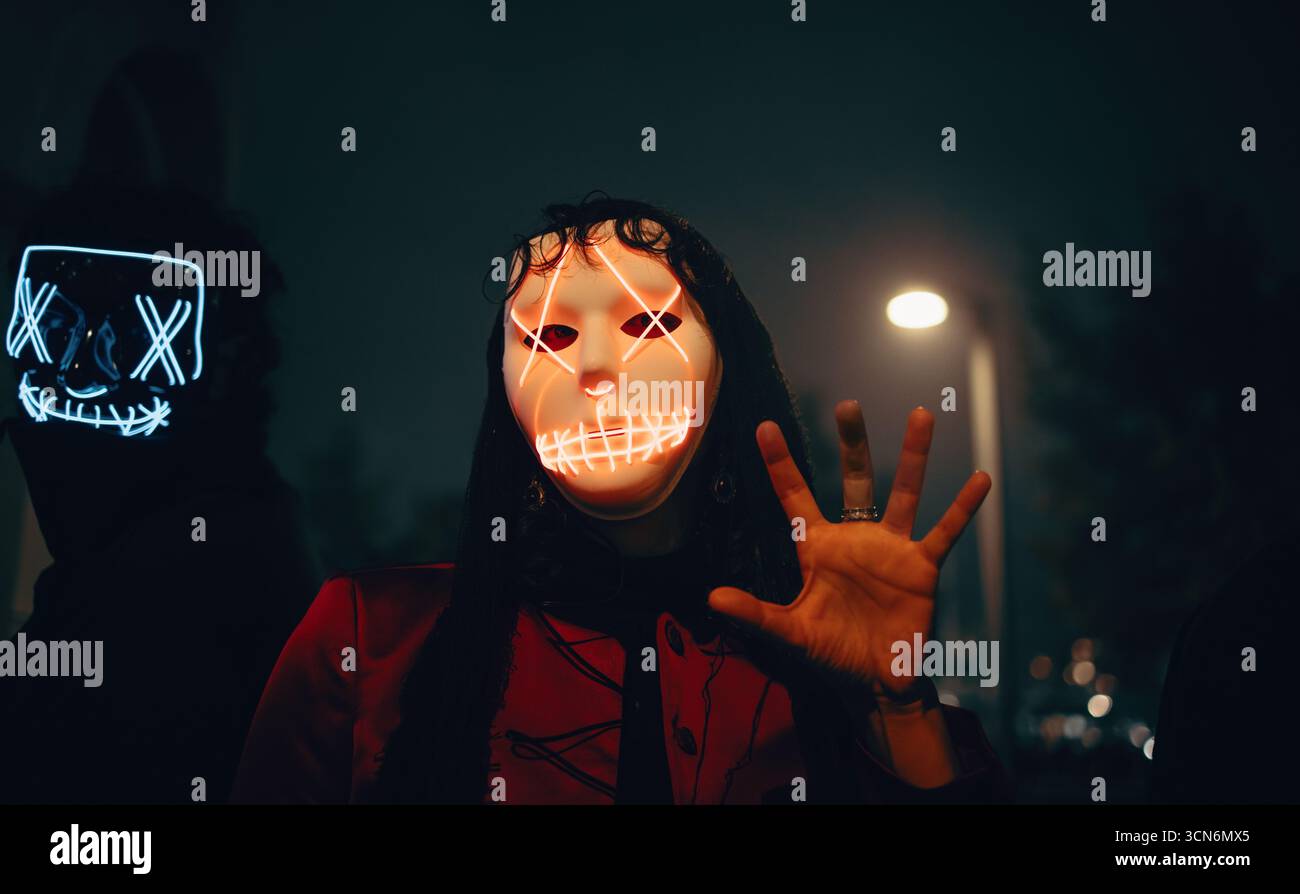 Le persone che indossano maschere illuminate si illuminano su uno sfondo urbano scuro, evocando mistero e intrighi. Foto Stock
