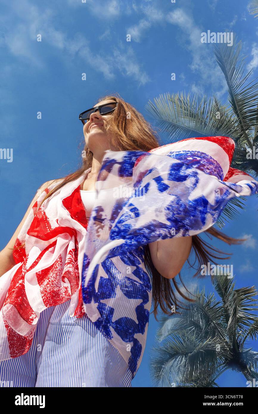 Una donna con occhiali da sole e una sciarpa americana in stile bandiera posa sotto alte palme in una giornata di sole. Sfondo tropicale, dinamica, patriottismo. Foto Stock