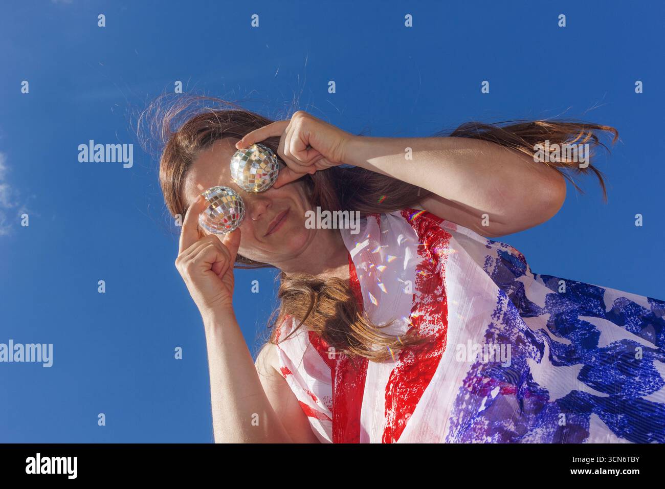 Una donna con una sciarpa in stile bandiera americana tiene due piccole palle da discoteca davanti a lei contro un cielo azzurro. Posa giocosa, atmosfera festosa e patriottica Foto Stock