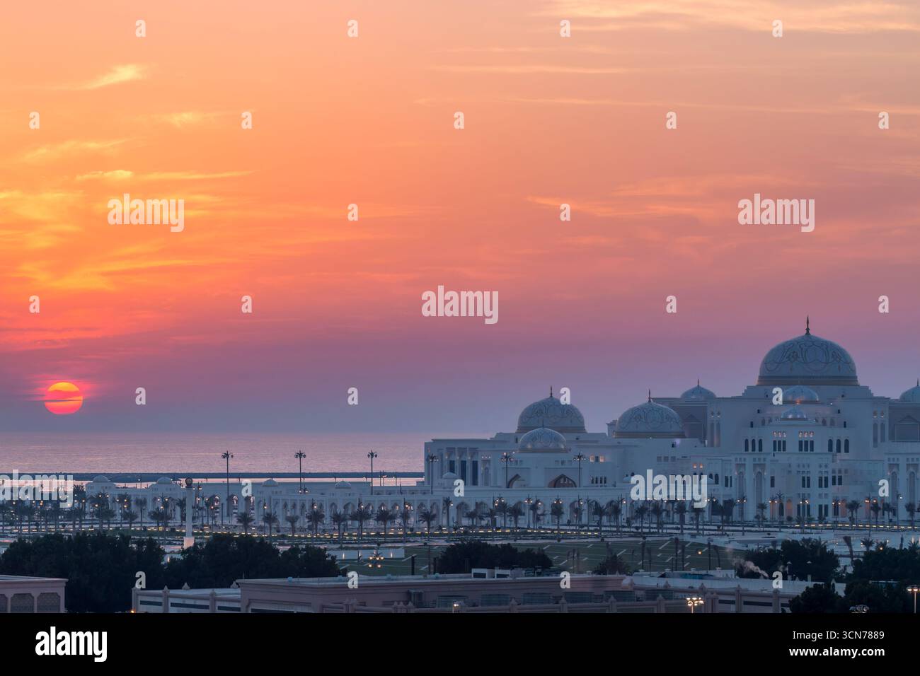 Emirati Arabi Uniti - Abu Dhabi - Qasr al Watan - il tramonto dipinge cupole e portici in una luce calda mentre il palazzo incornicia la costa oltre le palme Foto Stock