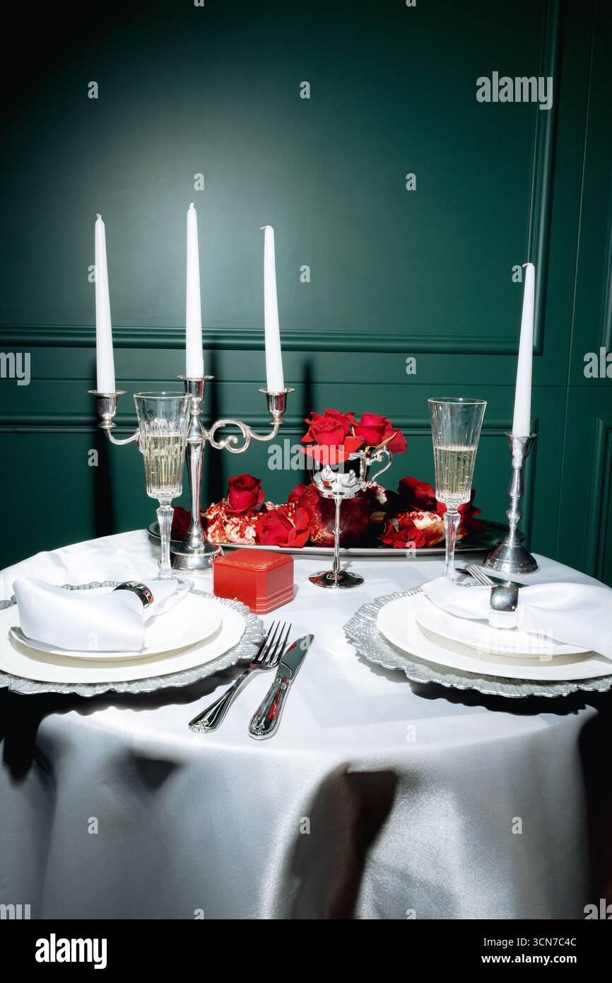 Un tavolo da pranzo splendidamente organizzato presenta candele tremolanti, bicchieri scintillanti e rose rosse fresche, creando un'atmosfera calda e intima. Foto Stock