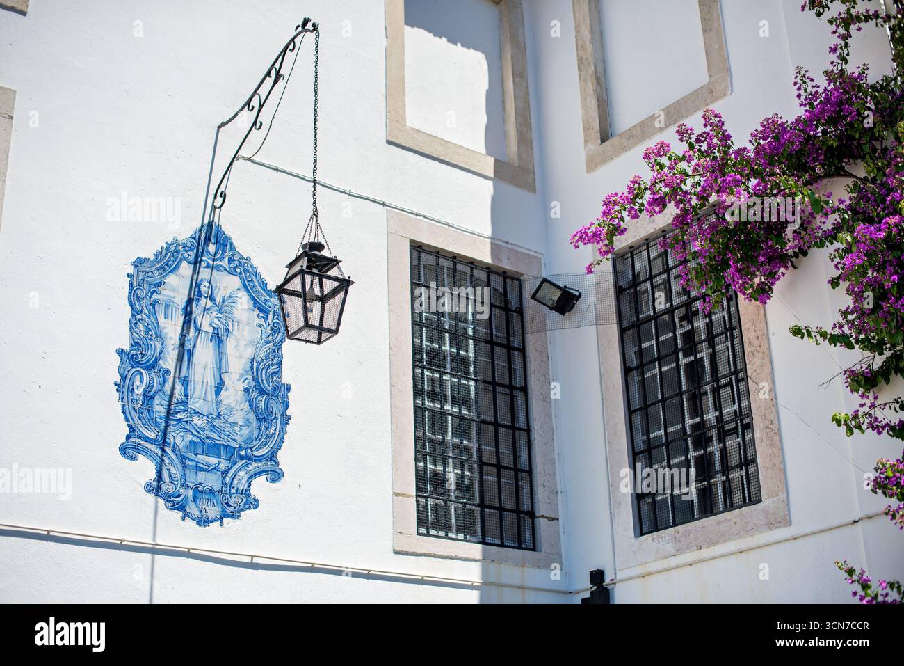 Mattonelle di Azulejo raffiguranti Santa Lucia Lisbona Portogallo // LISBONA, Portogallo - una piastrella azulejo raffigurante Santa Lucia è apposta su un muro bianco a Lisbona, Portogallo. Le piastrelle ornate blu e bianco presentano il santo che regge una fronda di palma e un libro, con scrolatura decorativa che circonda l'immagine centrale. Una staffa in ferro battuto si estende dal muro, reggendo una lanterna d'epoca. La facciata dell'edificio include finestre con griglie di metallo nero ed e' adornata da vibranti bouganville viola. Foto Stock