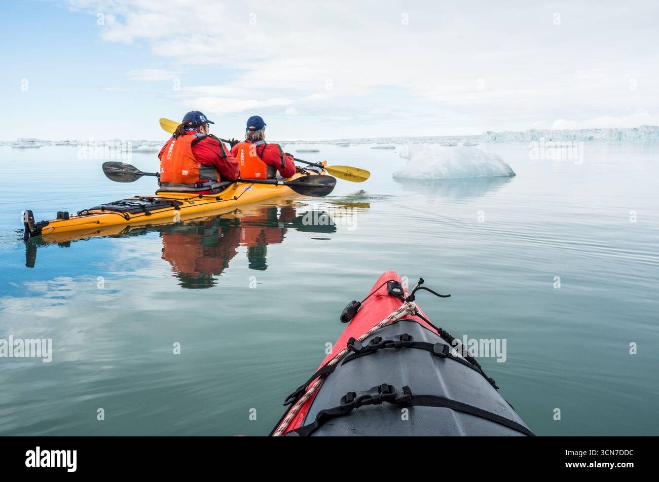 Kayakisti a Klerckbukta Nordaustlandet Svalbard Norvegia | NORDAUSTLANDET, Svalbard — Due kayakisti in giacche rosse remano tra iceberg alla deriva a Klerckbukta, mentre la linea pallida della fronte glaciale di Bråsvellbreen si estende attraverso la baia davanti a loro. Bråsvellbreen, un emissario della calotta glaciale di Austfonna, forma una delle più lunghe fronti glaciali continue dell'emisfero nord all'interno delle acque protette della riserva naturale di Nordaust-Svalbard. Foto Stock