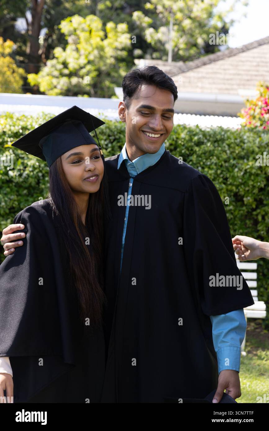 Amici indiani in piedi nel giardino del campus con abiti da laurea, mortaio vicino alla siepe Foto Stock