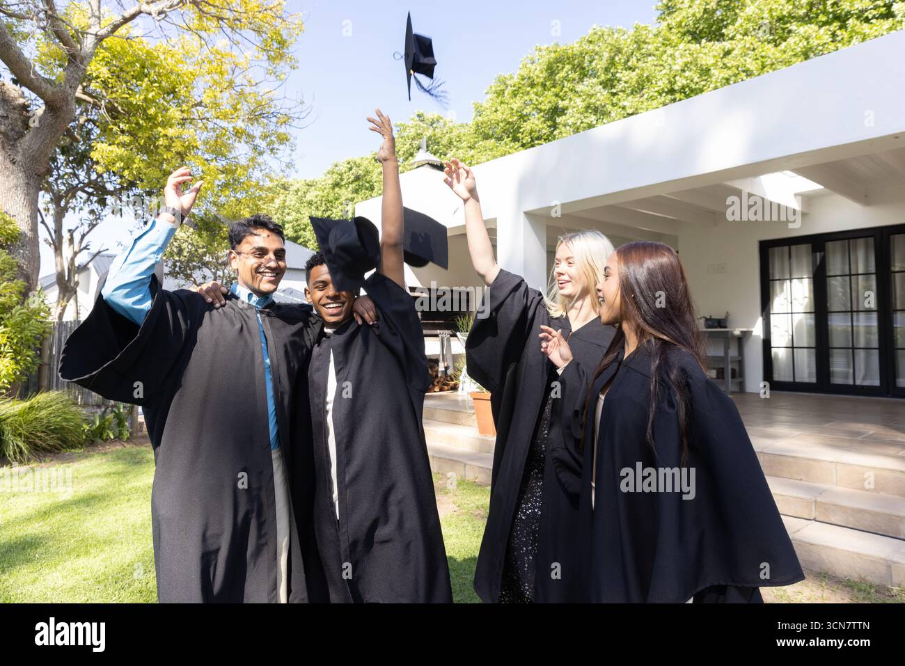 Diversi compagni di classe in camici accademici che lanciano cappellini per festeggiare la laurea sul prato Foto Stock