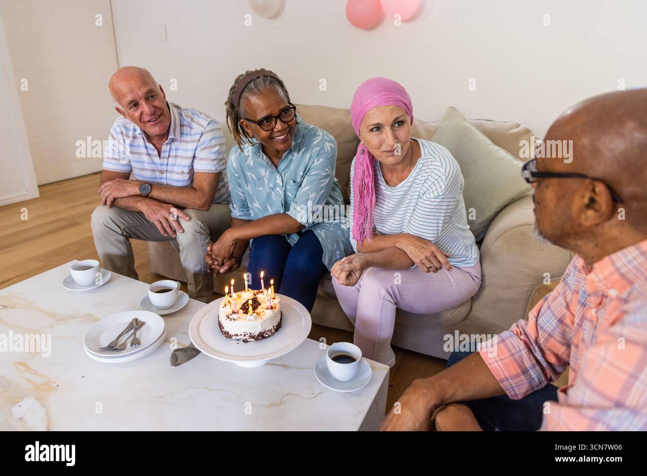 Diversi amici anziani festeggiano il compleanno a casa intorno al tavolino da caffè con torta e palloncini Foto Stock