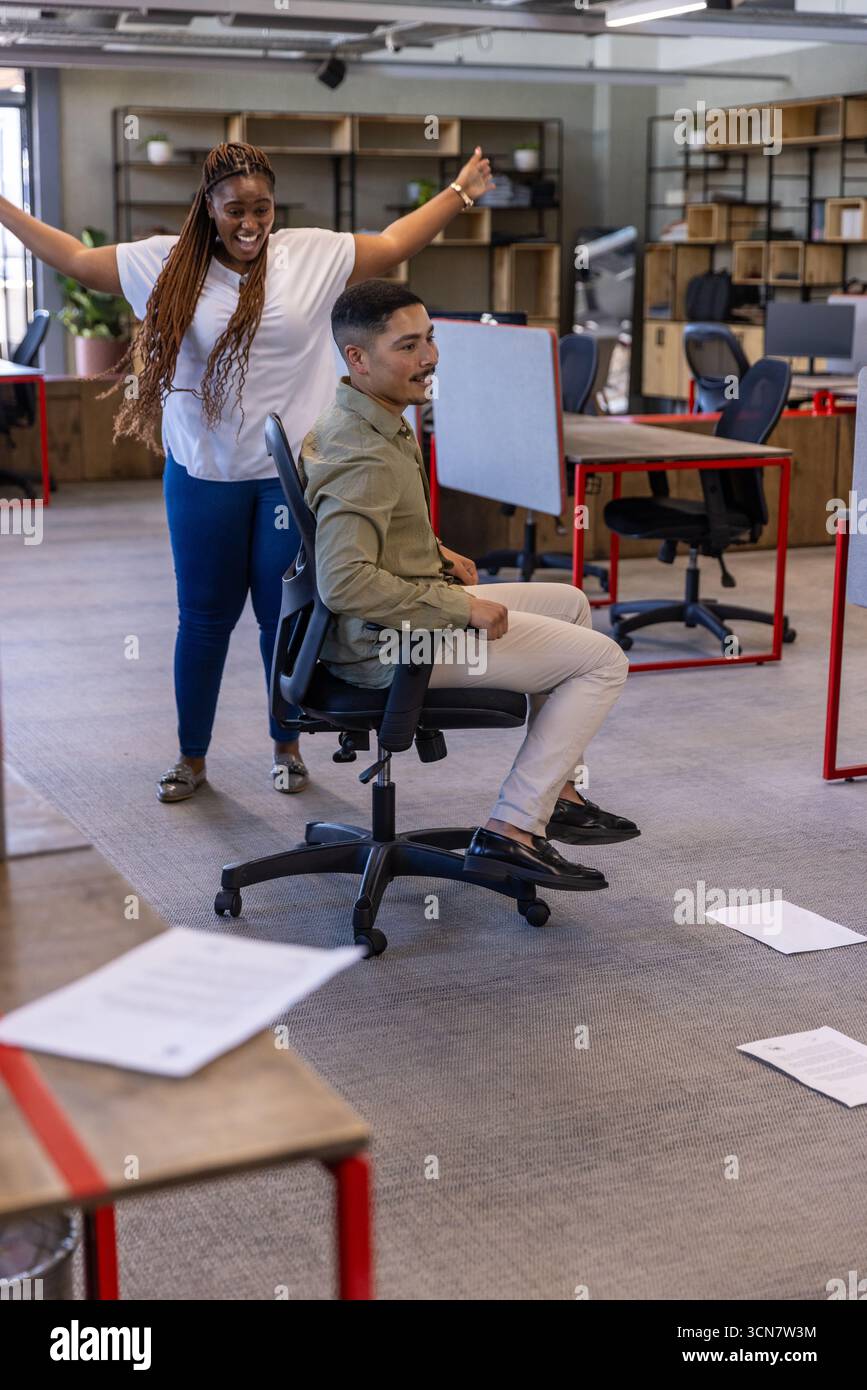 Diversi colleghi ridono mentre spingono il collega sulla sedia da ufficio in ufficio Foto Stock