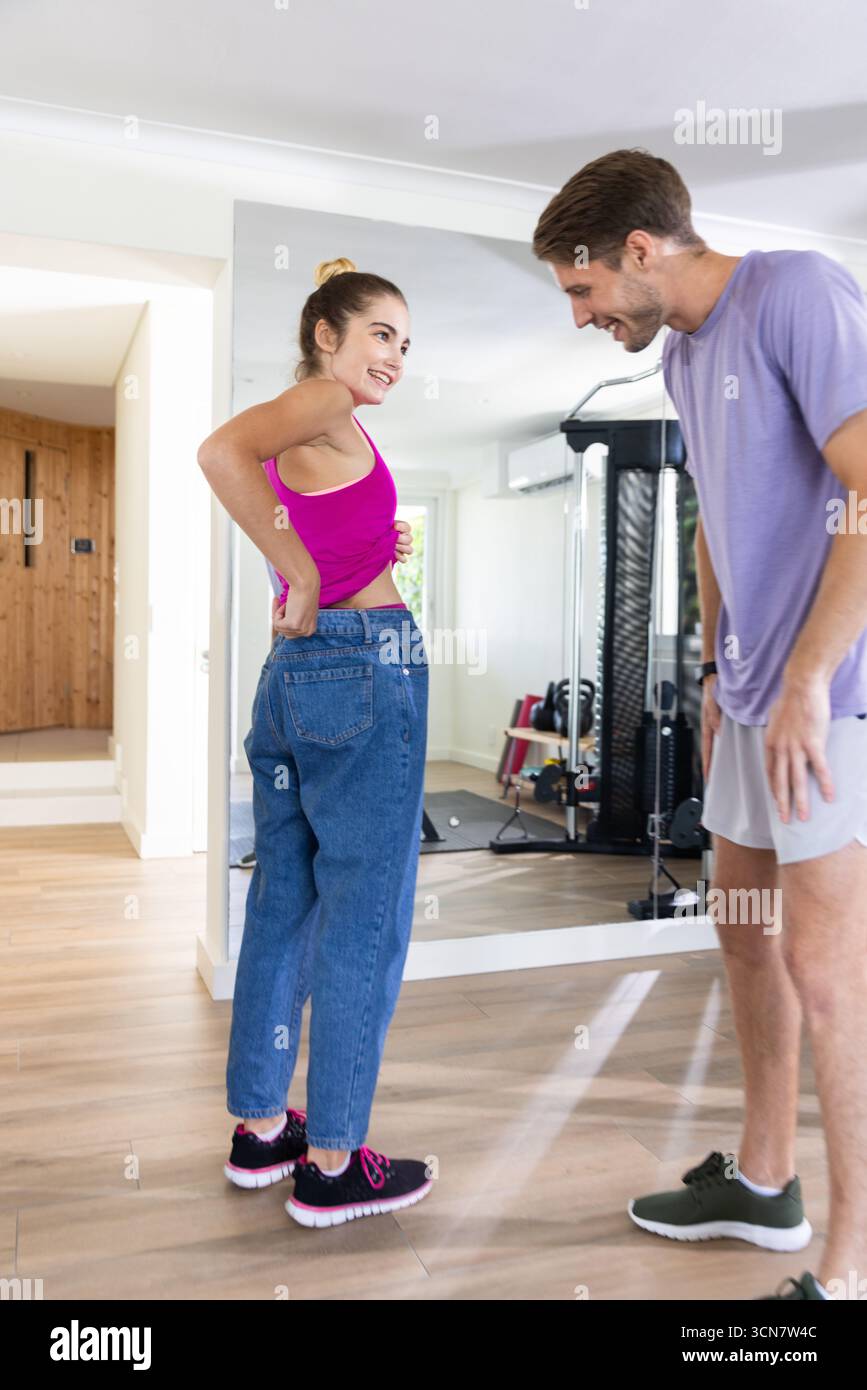 Abbina i Blue jeans sfusi mentre sorridi nella palestra di casa vicino allo specchio e alla macchina per sollevamento pesi Foto Stock